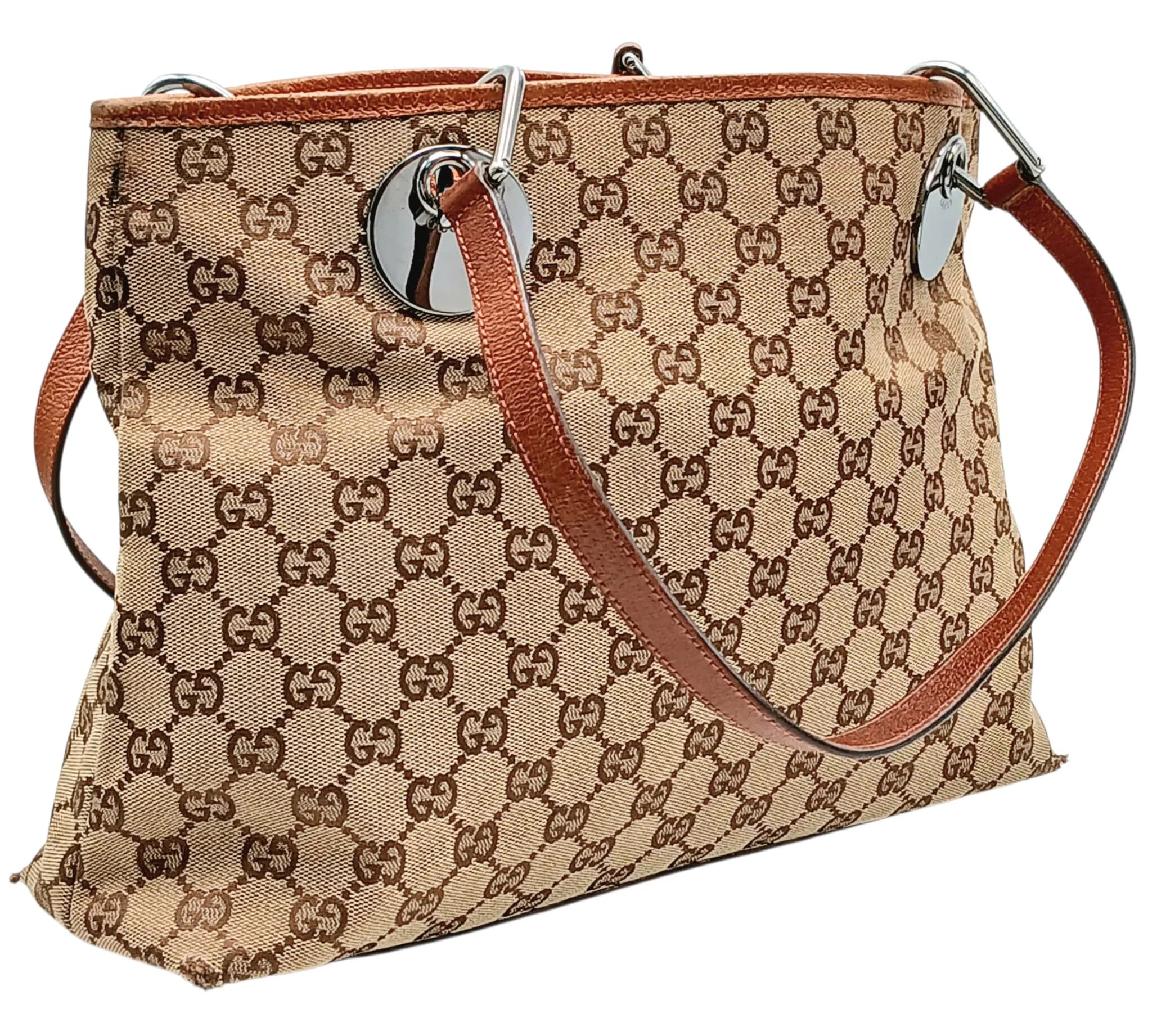 A Gucci GG Canvas Tote Bag. Beige and brown GG monogram canvas exterior with brown leather trim, silver-tone hardware, and double shoulder straps with circular Gucci-branded metal attachments. Open top to two main compartments with a central zipped compartment, brown textile-lined interior with a zipped inner side pocket and an open inner side pocket. Comes with a dust cover bag. Approximately 34cm length x 24cm height. In fair overall condition with wear to the base corners and white marks to the interior – please see photos or request a condition report. Ref: 27877